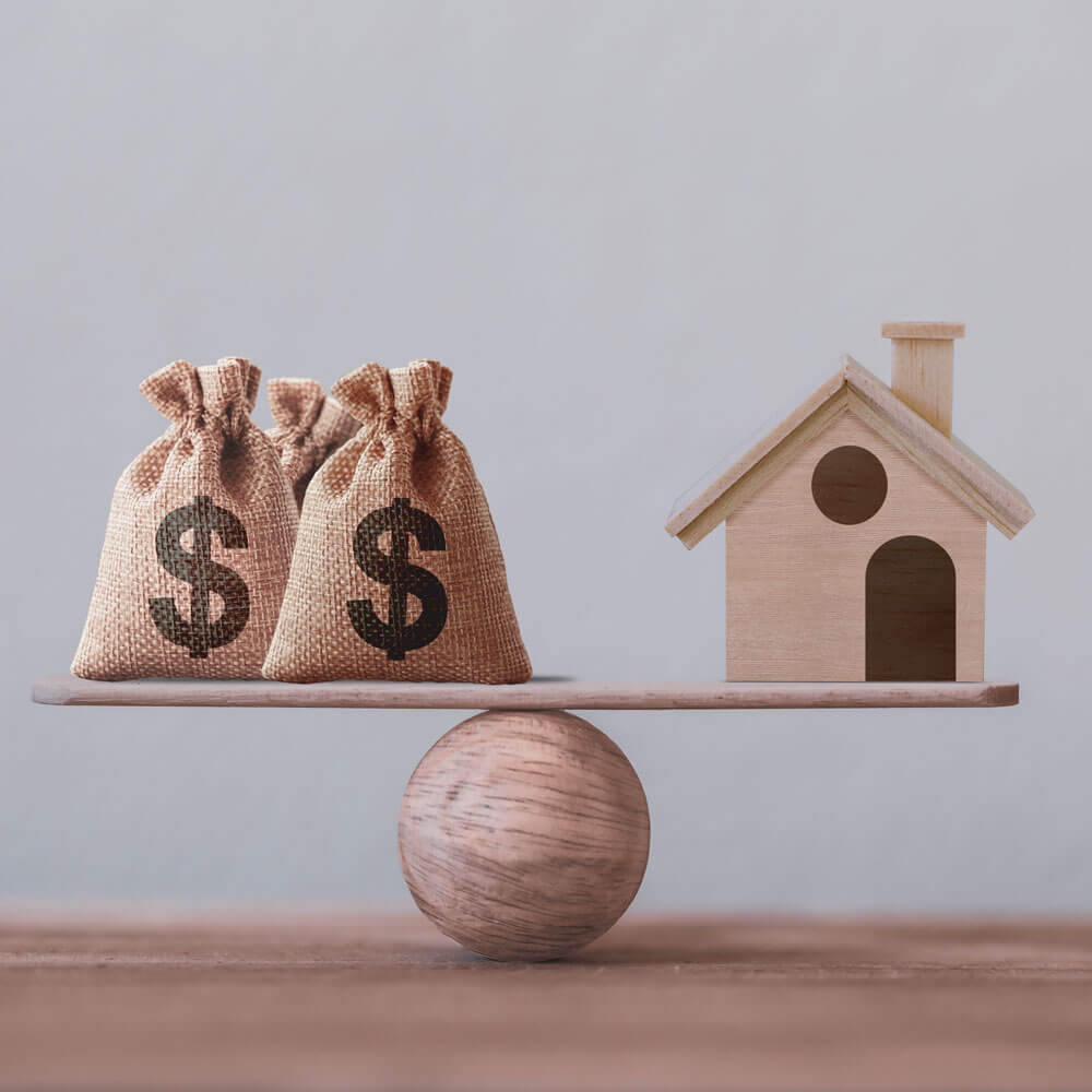 A balance scale with two bags of money on one side and a model house on the other side.